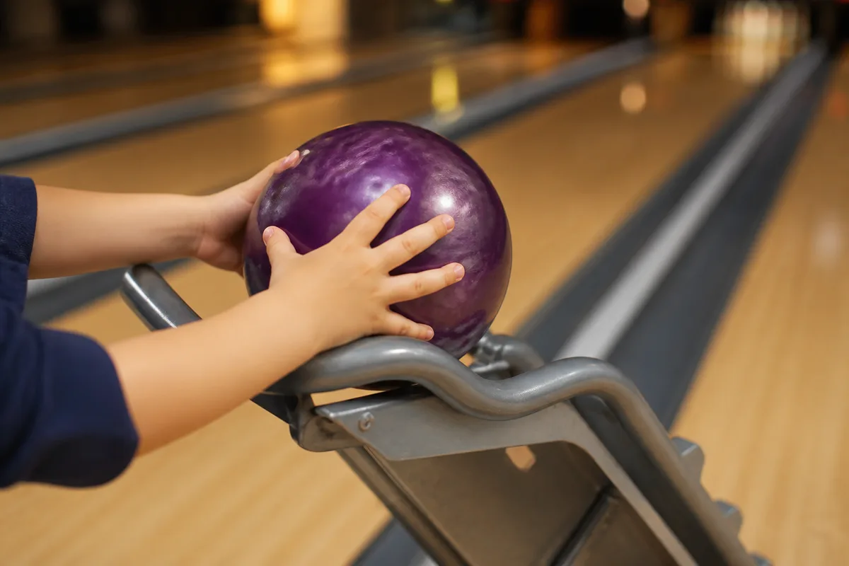 Un enfant pousse une boule de bowling posée sur une rampe métallique devant une piste éclairée