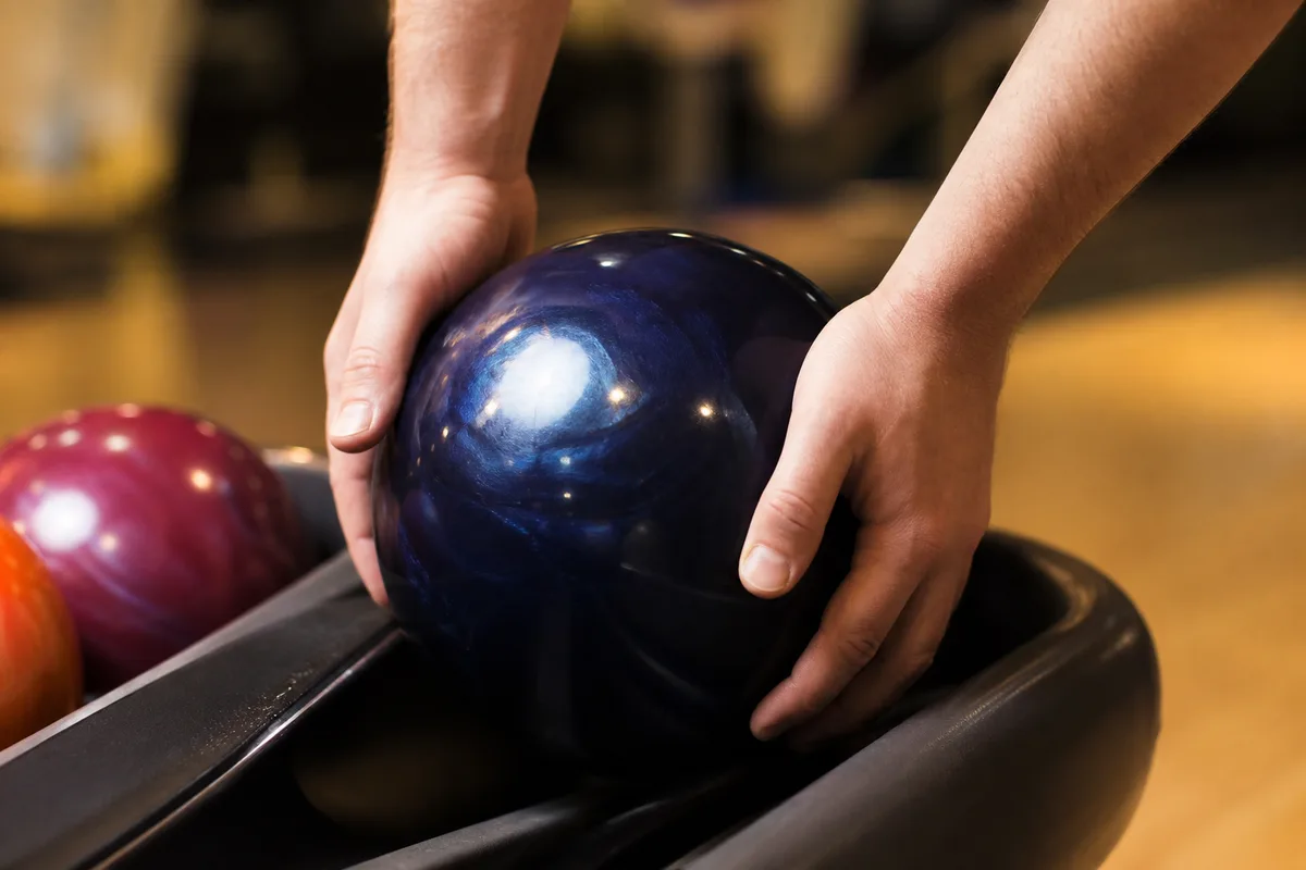 Mains d&rsquo;un joueur de dos saisissant une boule de bowling sur le retour de boules, éclairage chaud de salle